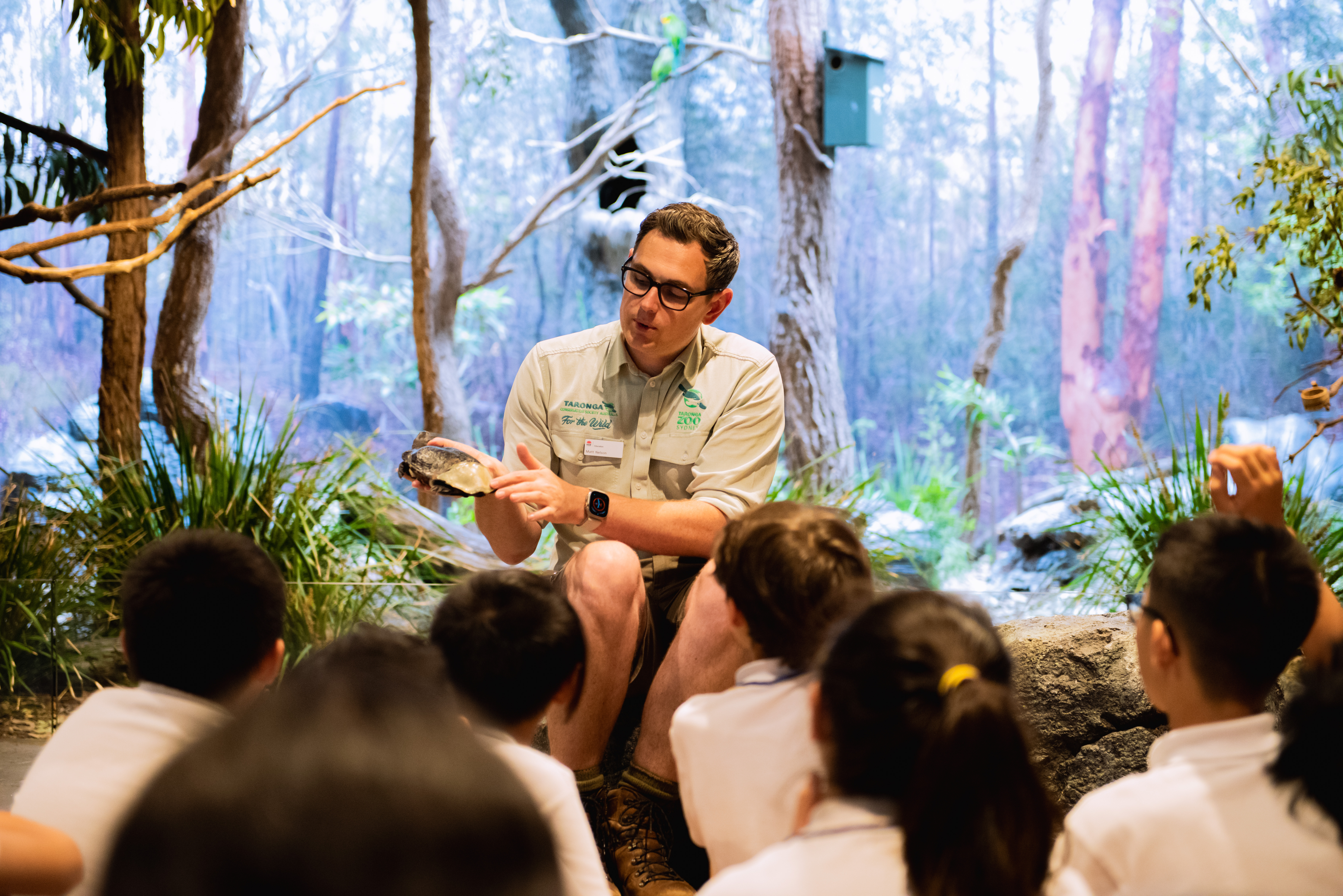 School students seated on the floor during a teacher‑led lesson in an immersive habitat classroom.