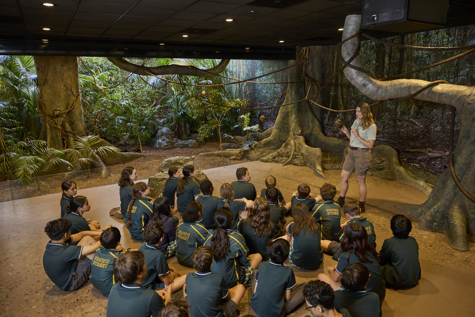Students seated indoors listening to a teacher in a rainforest-themed immersive classroom.