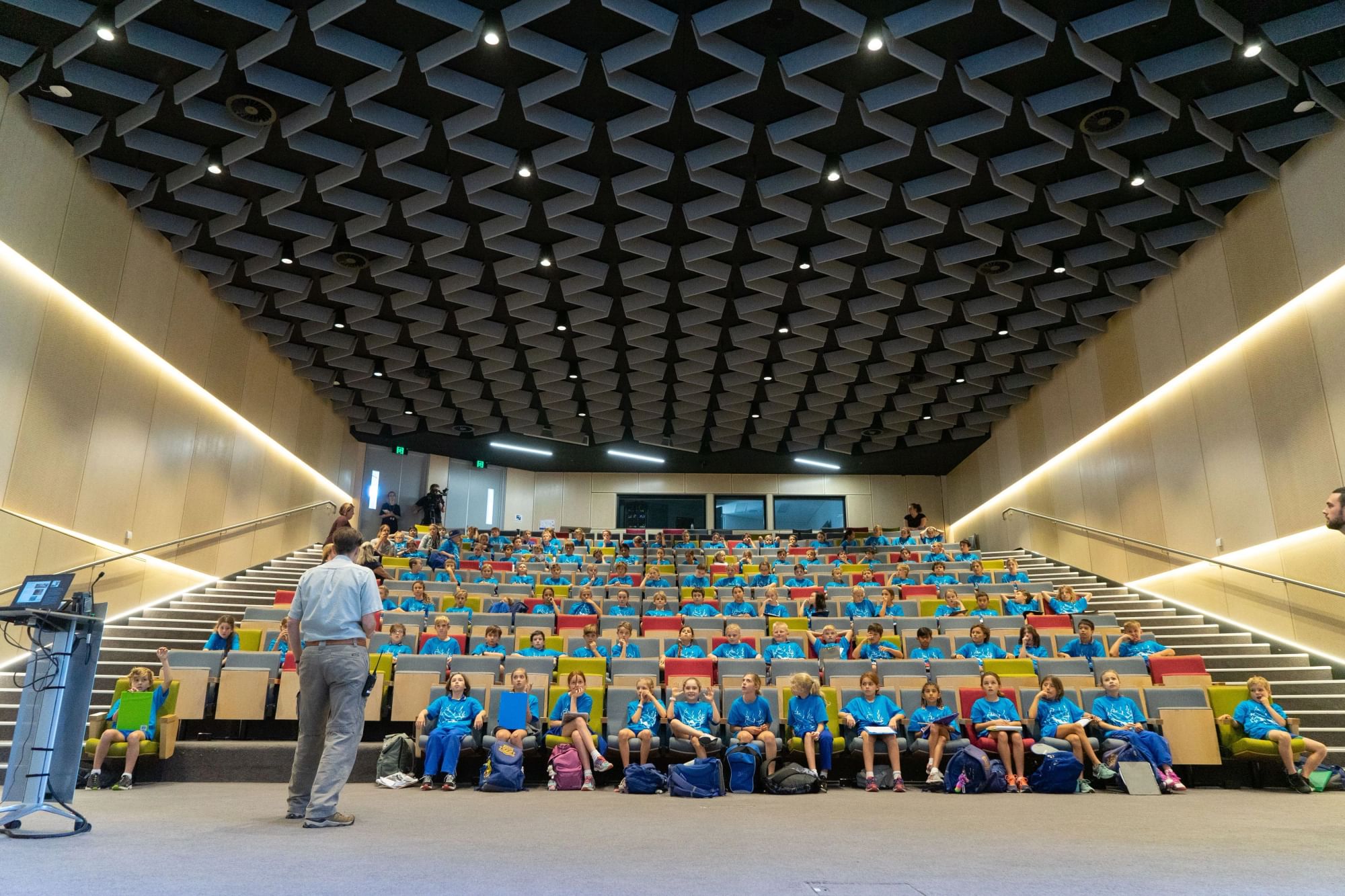 Students seated in a large auditorium listening to a teacher at the front.
